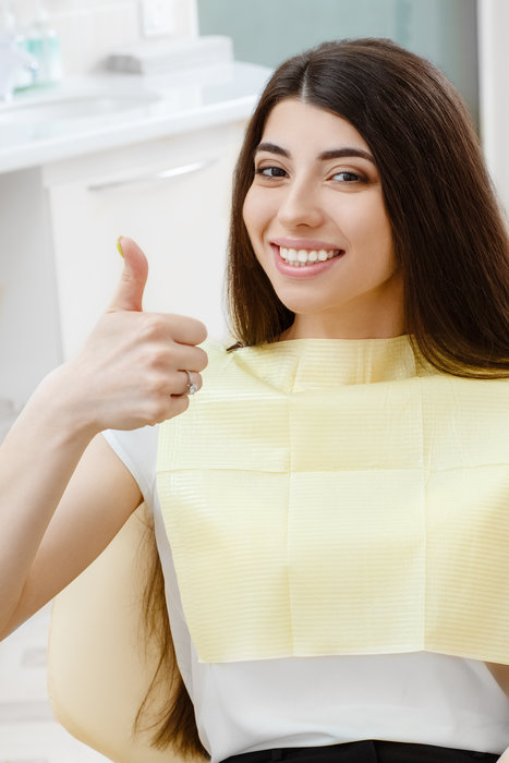 Showing her perfect smile. Beautiful brunette woman showing thumbs up smiling to the camera cheerfully with her perfect white and healthy smile sitting in a dental chair