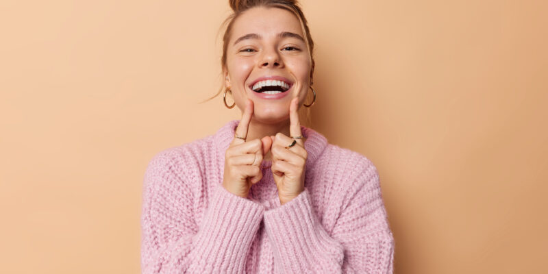 Positive young woman with combed hair points at her broad toothy smile feels very happy has white perfect teeth wears knitted sweater isolated over beige background. People and emotions concept
