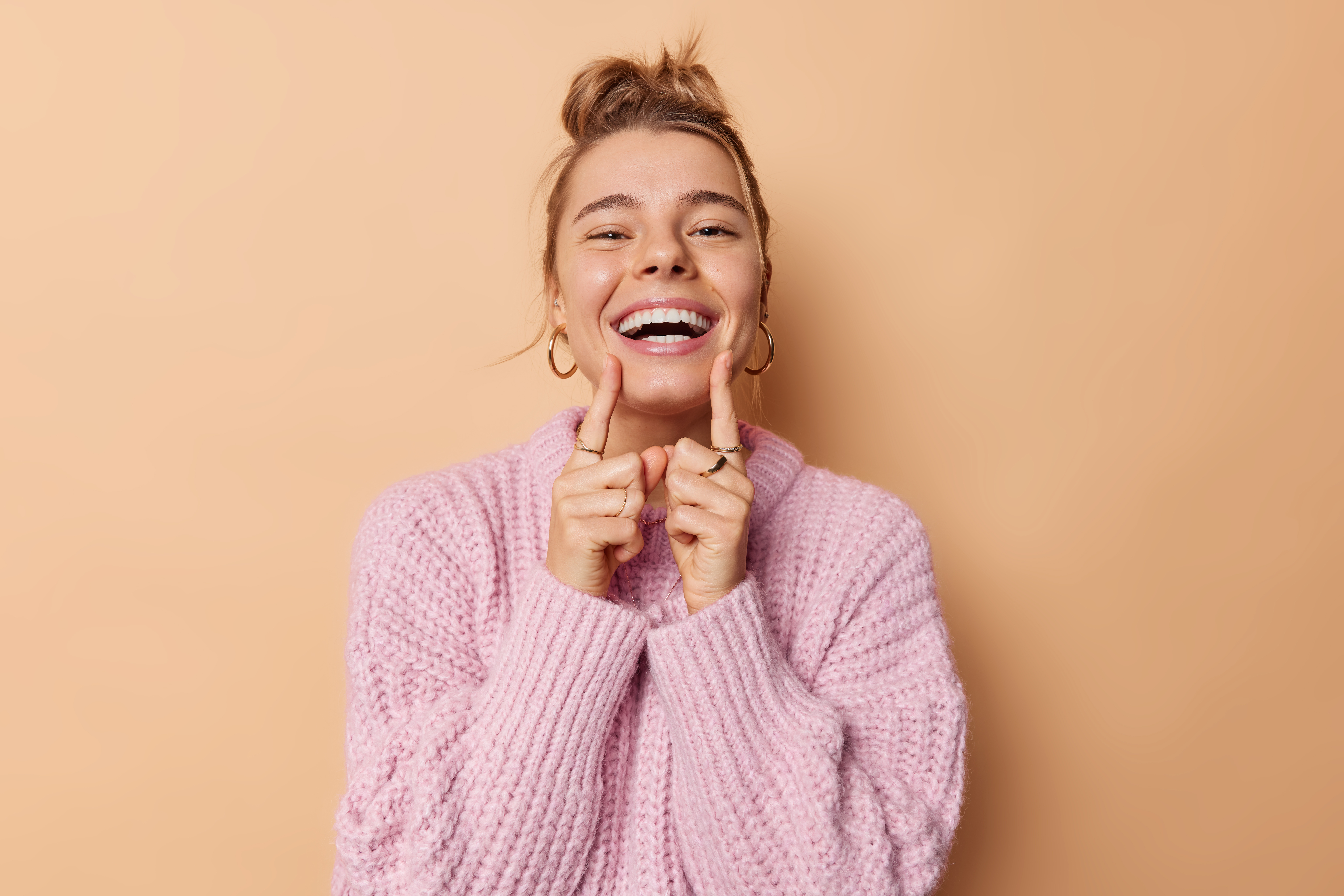 Positive young woman with combed hair points at her broad toothy smile feels very happy has white perfect teeth wears knitted sweater isolated over beige background. People and emotions concept