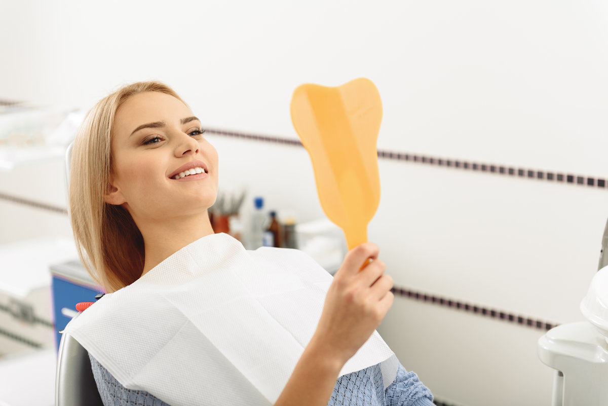 Attractive joyful woman looking at mirror while sitting on dental chair in stomatological room. She admiring her white smile