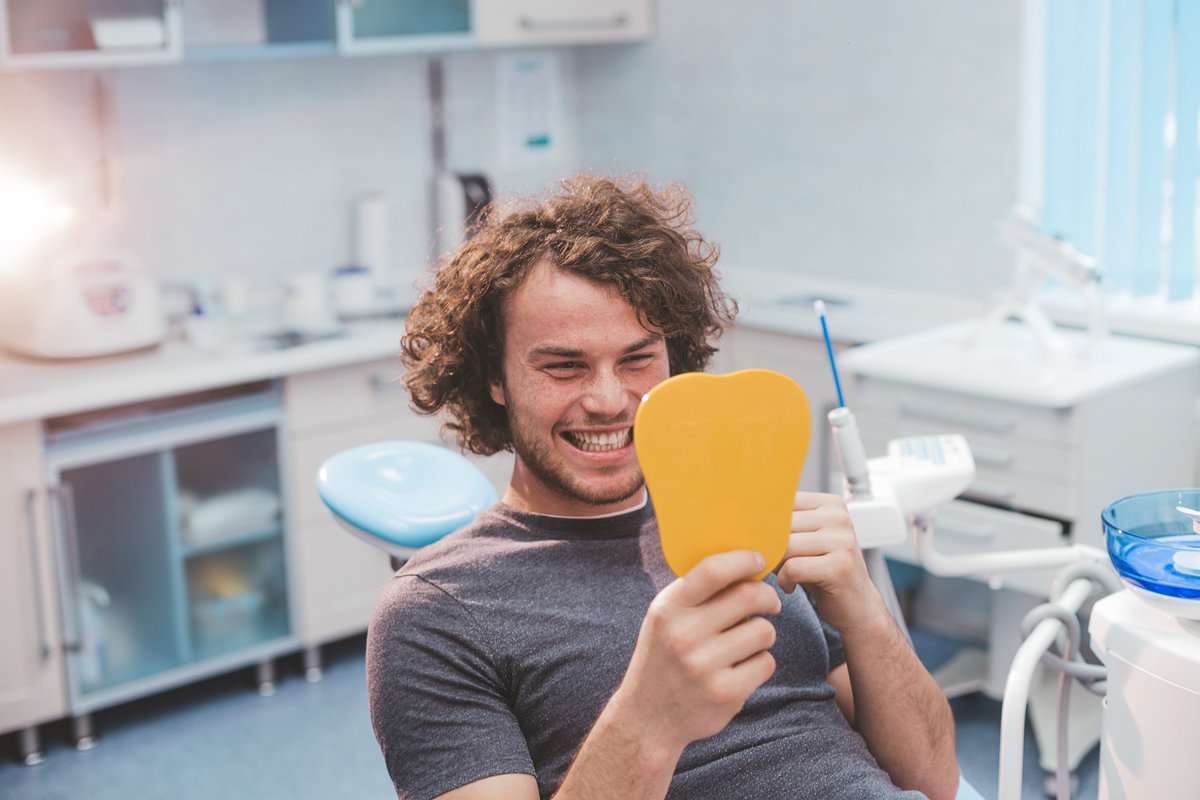 Charismatic smiling young man on the dentist chair , after a oral hygiene procedure take a mirror and looking at his white healthy teeth and are very happy for results