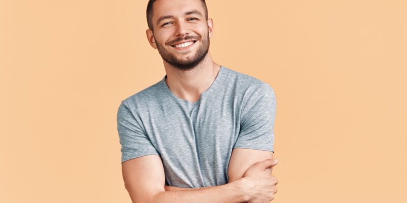 Happy smiling handsome man with crossed arms looking to camera over beige background. Emotions concept