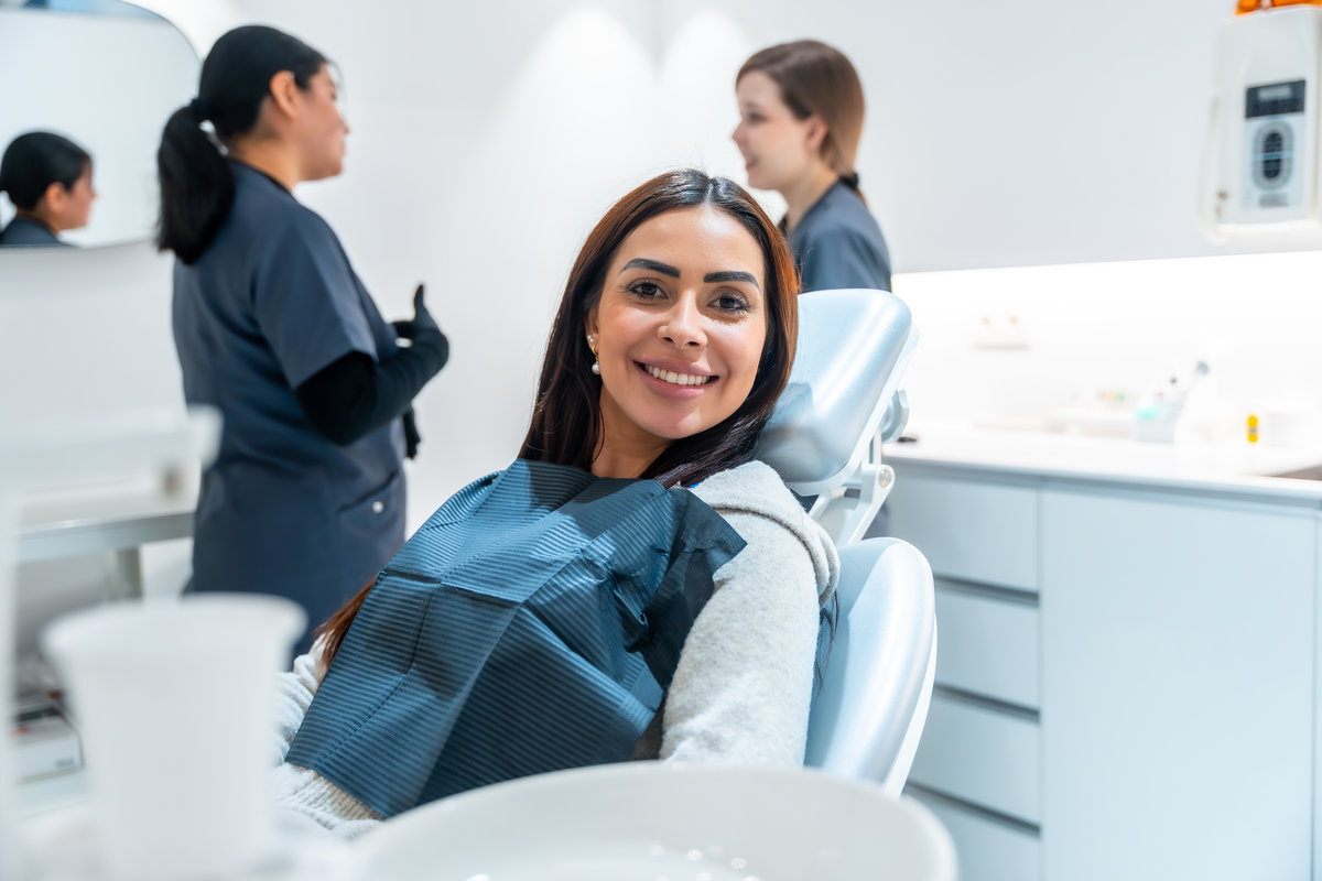 Young woman smiling while sitting on dentist chair during medical checkup, with two dentists talking in the background