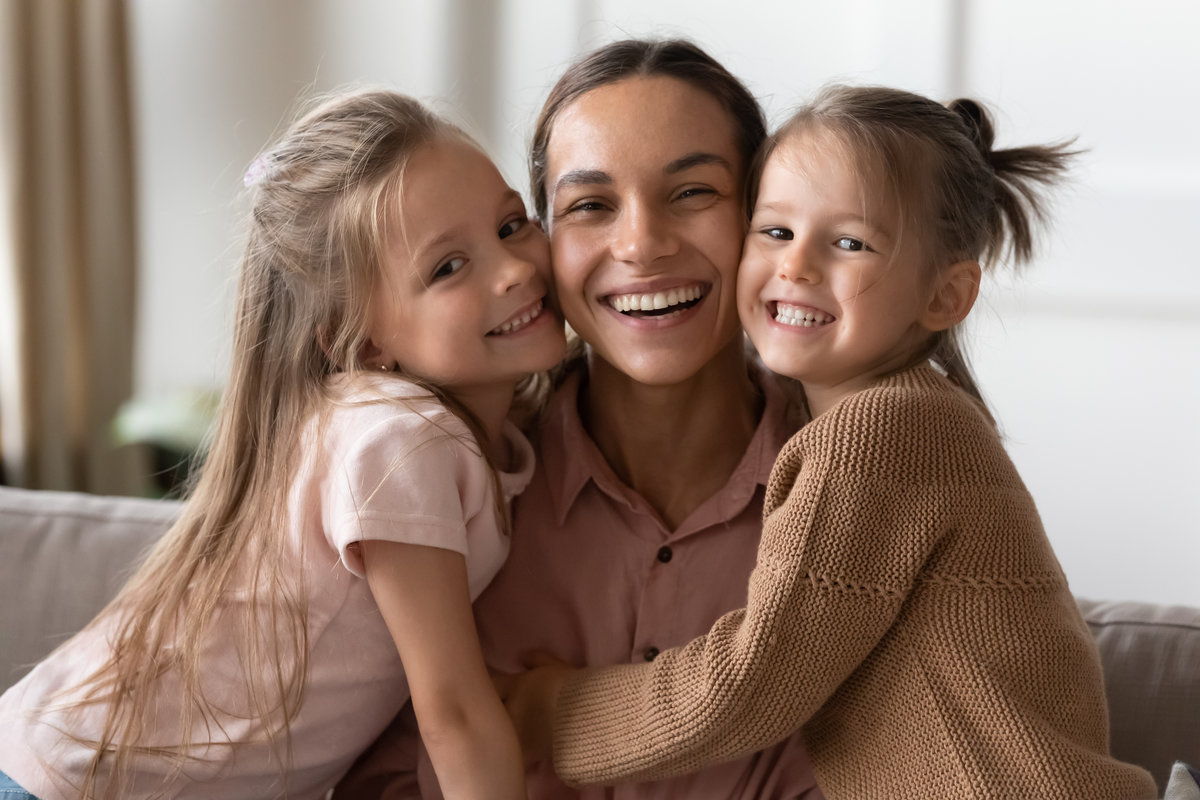 Beautiful female family young smiling single mother and cute little children daughters embracing looking at camera, funny small kids girls hugging happy mum bonding sit on sofa together, portrait