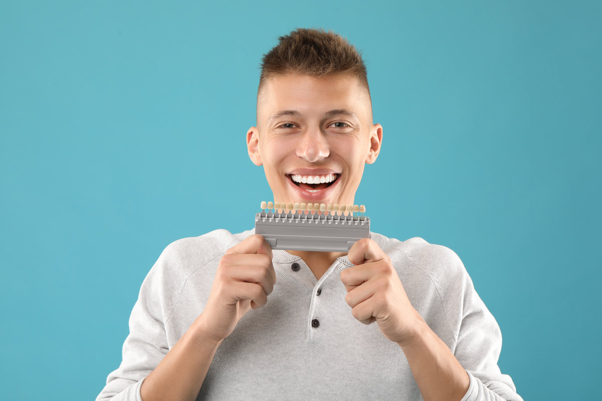 Happy young man with teeth color samples on light blue background. Dental veneers