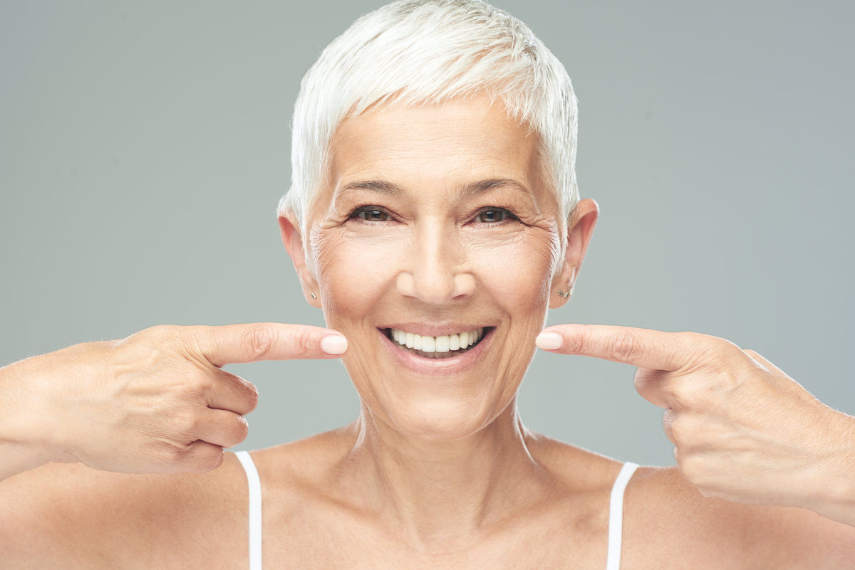 Beautiful Caucasian smiling senior woman with short grey hair pointing at her teeth and looking at camera. Beauty photography.