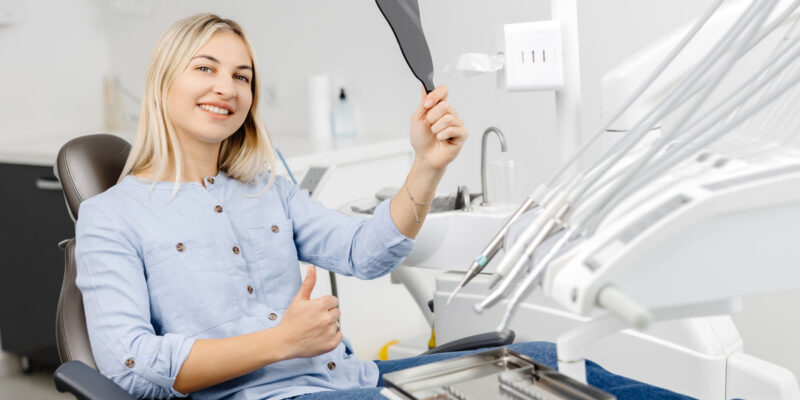 Smiling patient is checking her teeth after dental procedure using small mirror in modern dental clinic