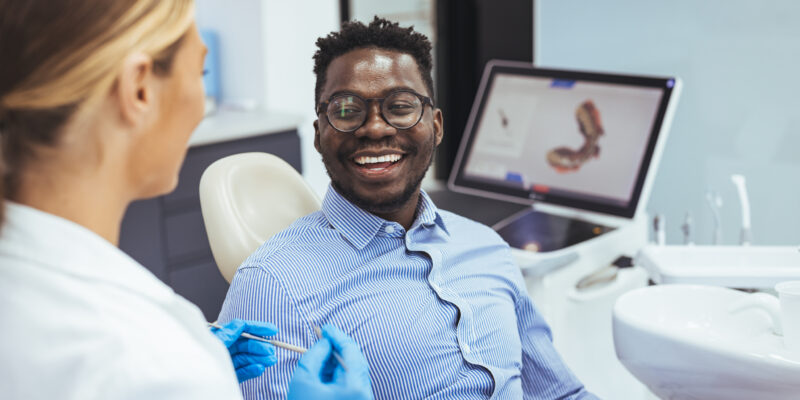 Smiling african guy in dentist chair looking with trust at his doctor, close up. Young African American Man Getting Teeth Treatment With Professional Stomatologist At Modern Clinic, Closeup